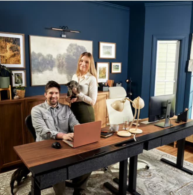 Two people in a blue office room with a desk and monitors.