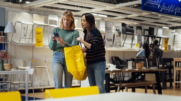 Two people in a furniture store holding a yellow shopping bag and checking a phone near dining tables and chairs.