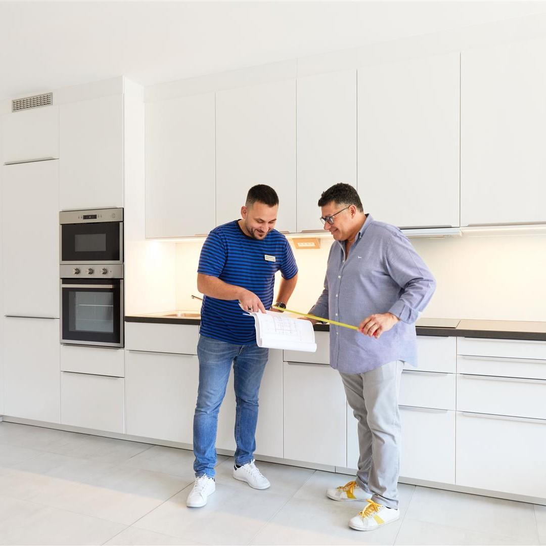 Two people in a modern kitchen; one holding a measuring tape, the other a clipboard. White cabinets, built-in oven and microwave.