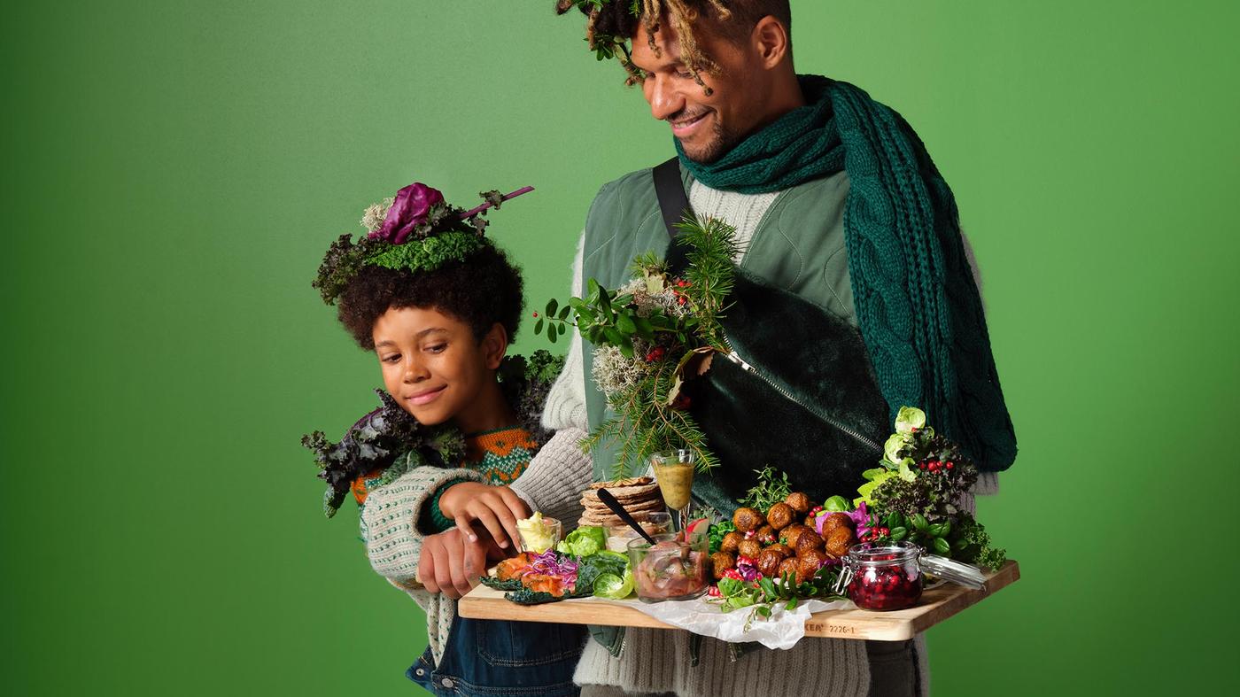 Two people in green-themed outfits holding a wooden tray with assorted vegetables and festive dishes.