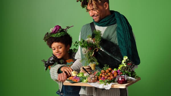 Two people in green-themed outfits holding a wooden tray with assorted vegetables and festive dishes.