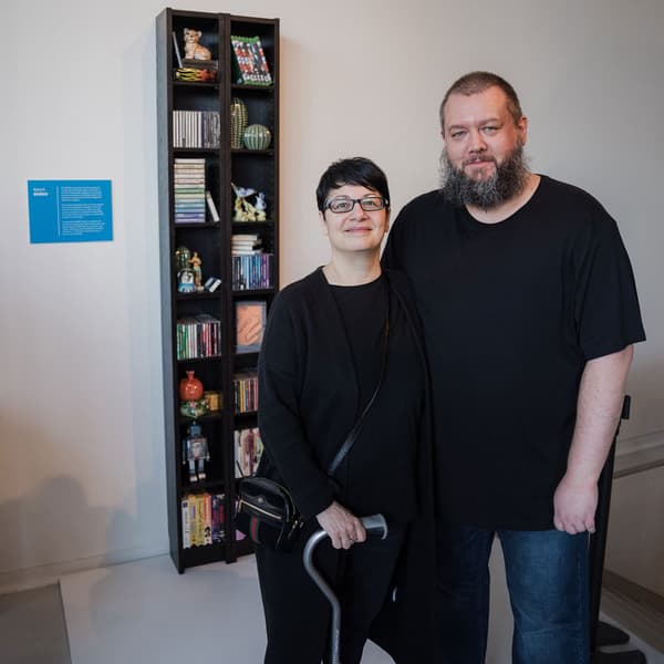 Two people pose in from of an IKEA BENNO bookcase, holding different items such as decorations, books, toys, and gaming CDs.