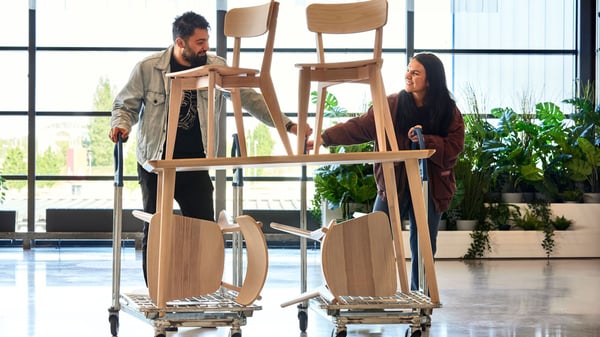 Two people push carts stacked with wooden chairs inside a bright, modern space with large windows and plants.