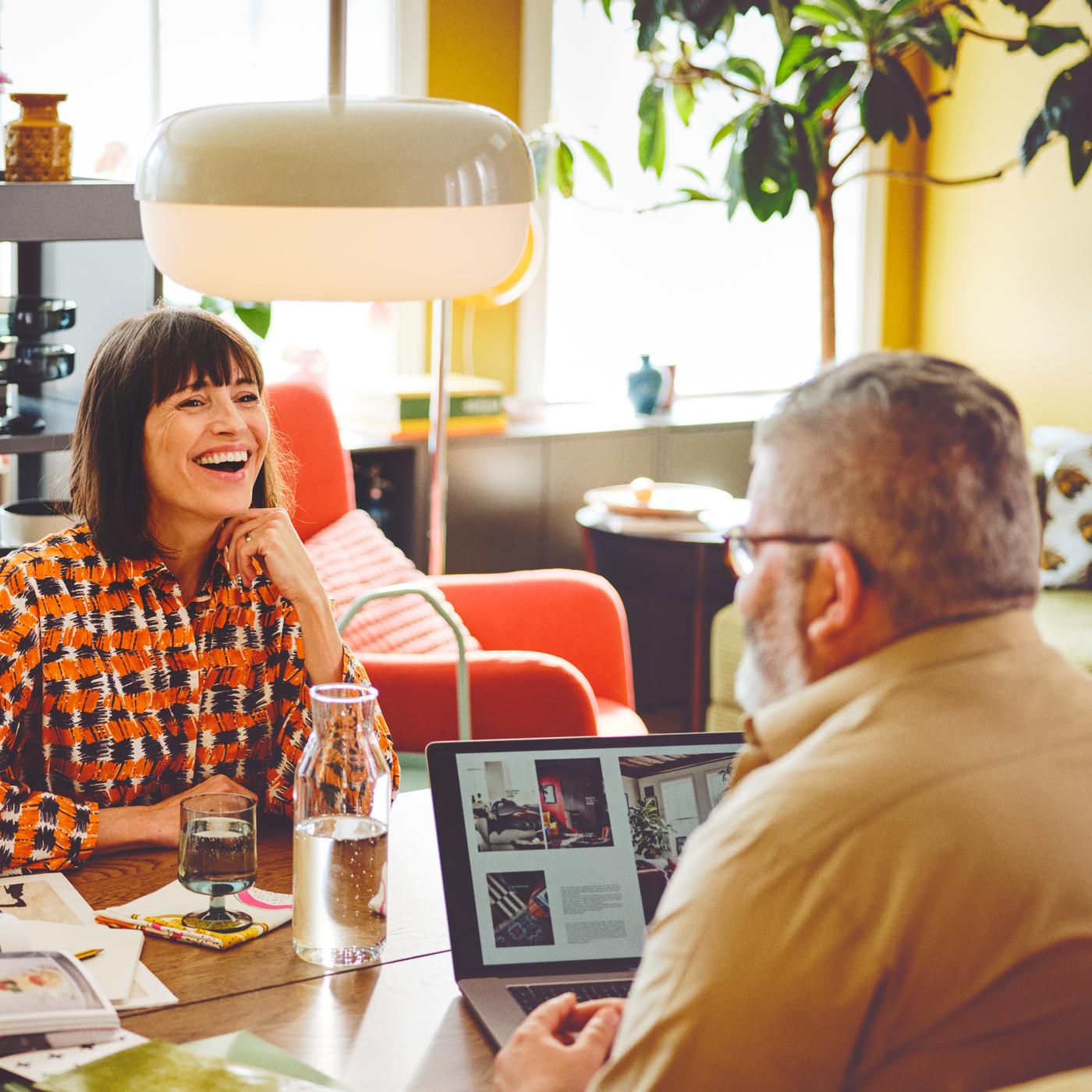 Two people sit across a table in a bright room. A laptop and carafe sit on the table. A BLÅSVERK pendant lamp hangs down.