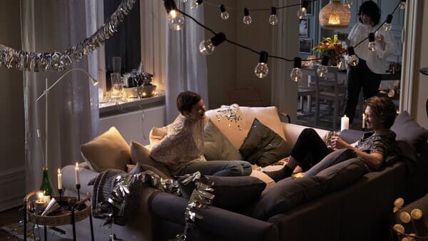 Two people sit together in two sofas that have been pushed together in the middle of a room, laughing and chatting.