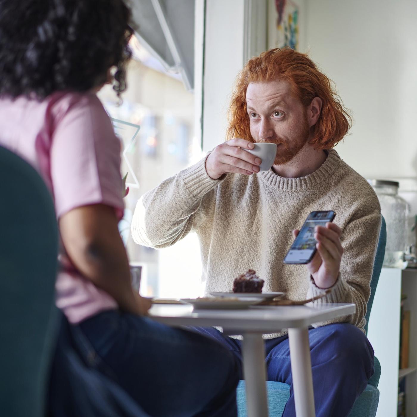 Two people sitting at a table, one drinking an espresso and showing the other something in the IKEA app.