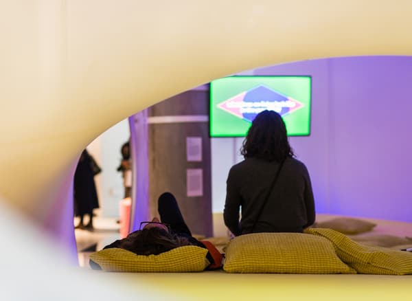 Two people sitting on cushions watching a film in an exhibition space.