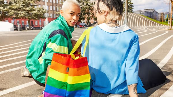 Two people sitting on the ground with a STORSTOMMA rainbow bag on their arm.