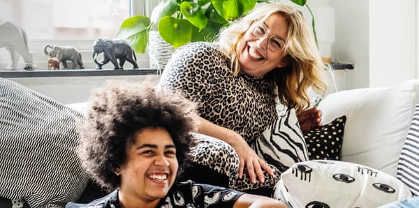 Two people smiling lounging on a sofa with beige and black cushions, a plant and window in the background.