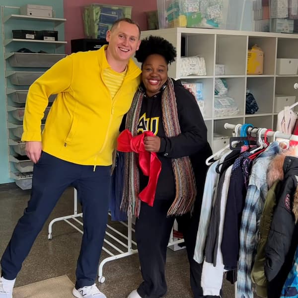 Two people smiling next to a clothing rack and one person holding a stuffed heart-shaped toy.