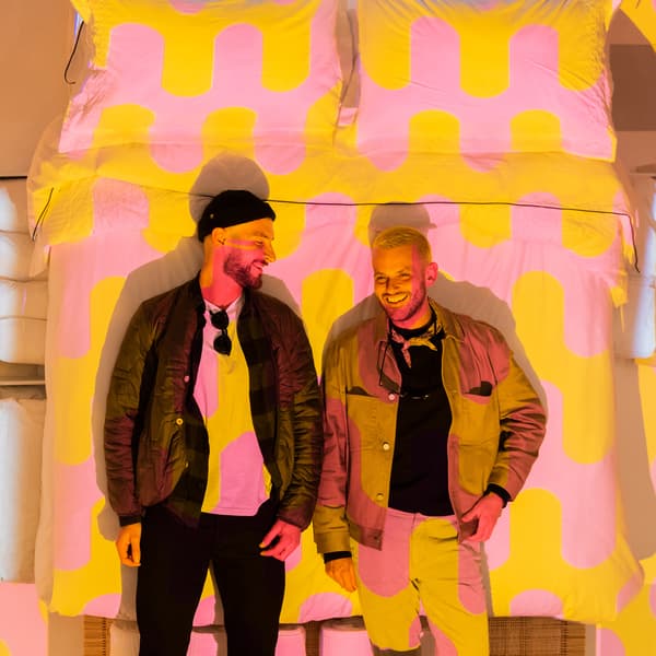 Two people stand in front of an image of a bed with graphic pink and yellow bedding.