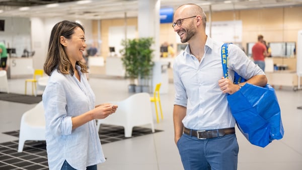 Two people talking in an IKEA store lobby with white chairs and a large blue shopping bag.