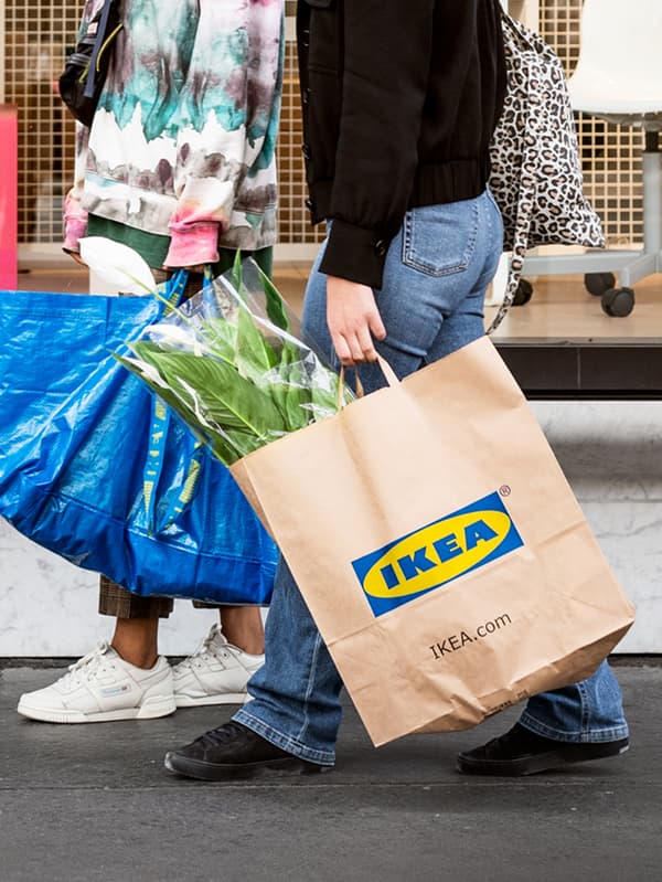 Two people who have been shopping at IKEA, one carrying a blue FRAKTA bag and the other an IKEA paper bag containing plants.