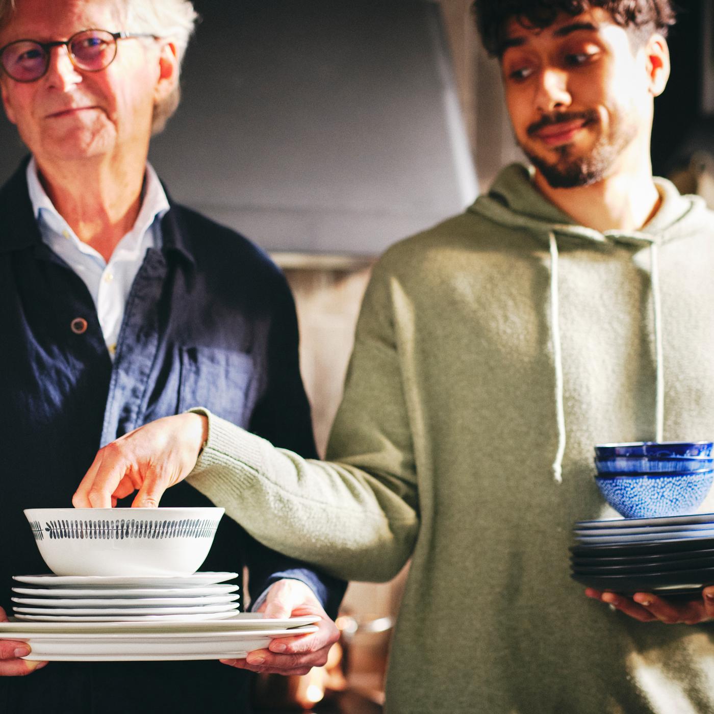 Two people with one of them holding a stack of white UPPLAGA serving and side plates and a white/patterned FRIKOSTIG bowl.