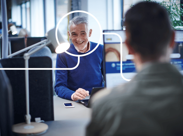 Two people working at desks in a modern office with computer screens, lamps, and partitions between workstations.