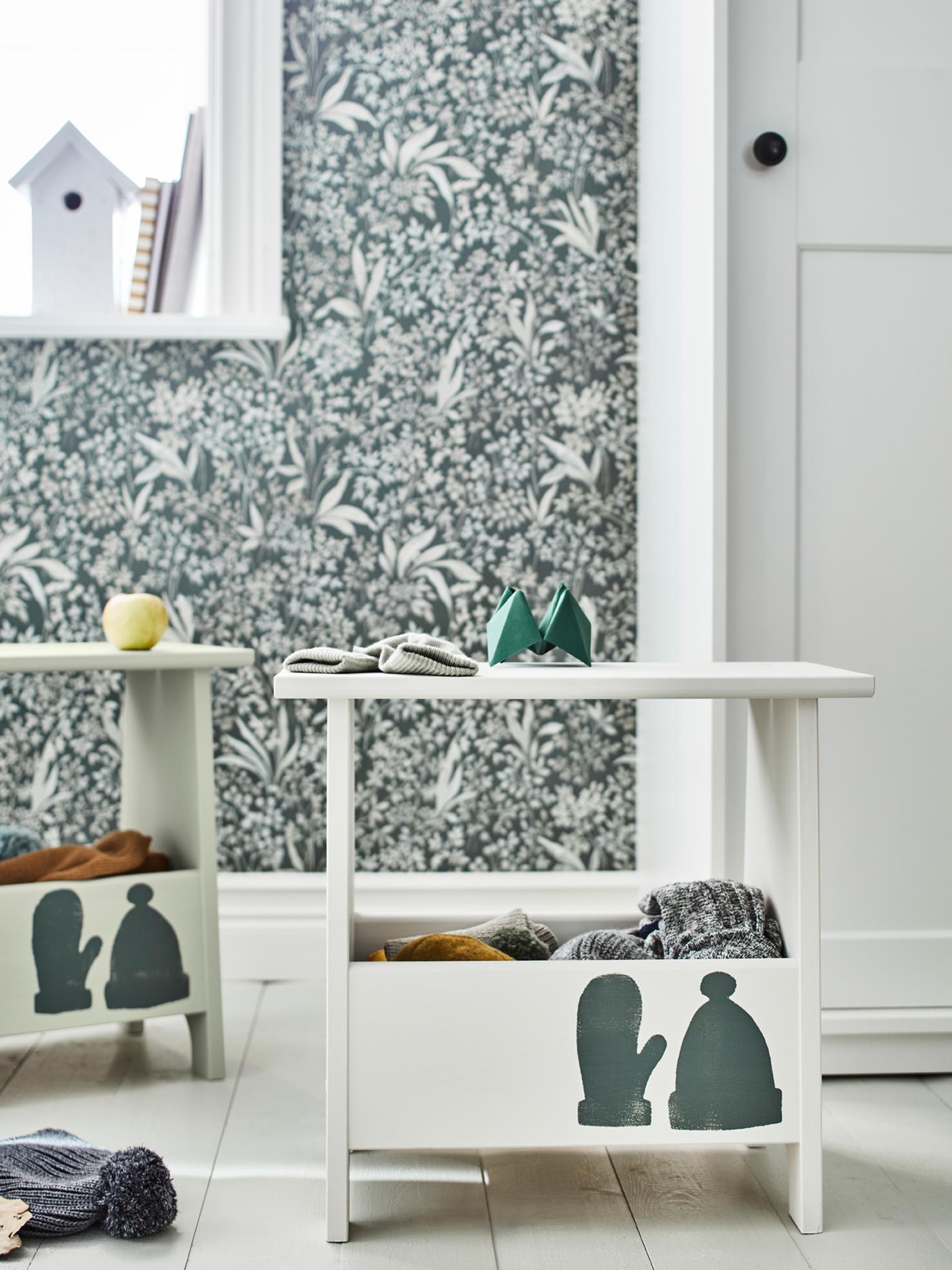 Two PERJOHAN stools with storage, painted white and with a hat and a mitten painted on them, stand in a hallway.