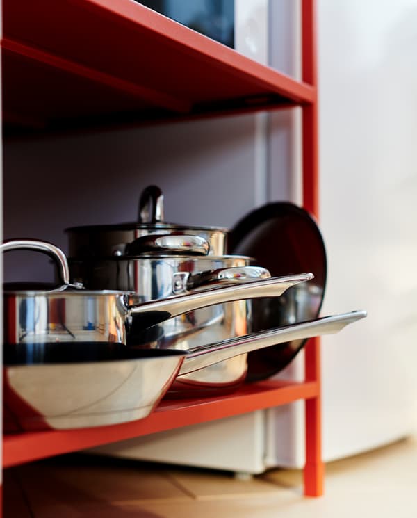 Two pots, one saucepan, one frying pan and lids in stainless steel are stored on the bottom shelf on a red shelving unit.