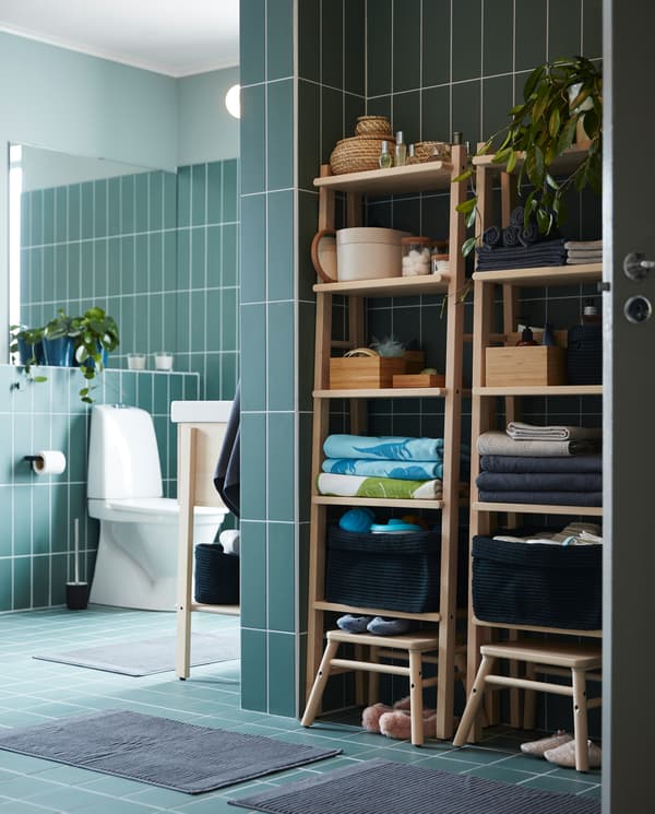 Two shelving units in birch with towels, baskets and boxes on the shelves, two birch step stools and dark grey bath mats.