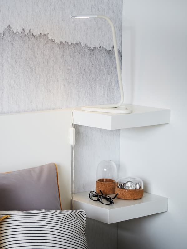 Two small, white IKEA LACK shelves mounted in a corner and used as a bedside table with lamp and decorative items.