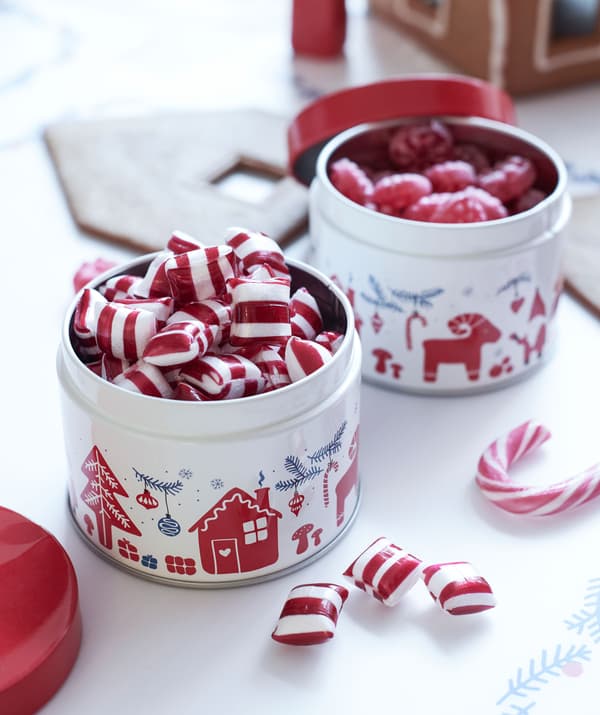 Two small Yuletide-patterned tins filled with hard candy standing on a surface next to a gingerbread-house construction site.