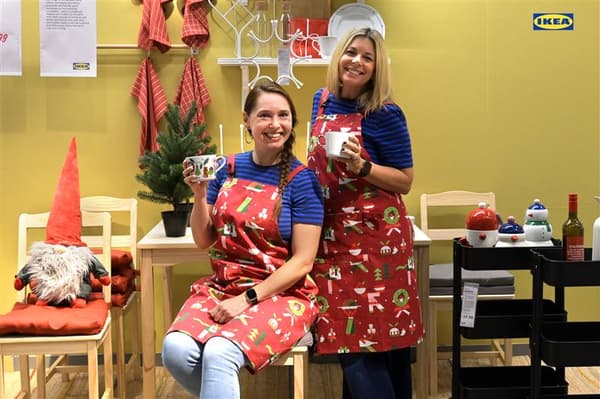 Two smiling women in matching blue striped shirts and red holiday aprons holding mugs in a festive dining display setup.