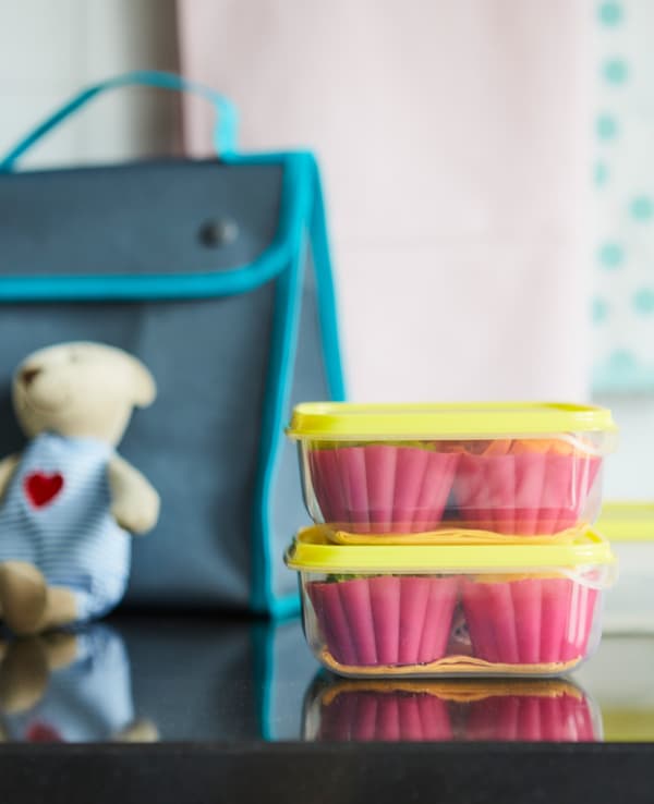 Two stacked glass food containers with yellow lids, containing pink muffin cups, near a school bag on a worktop.