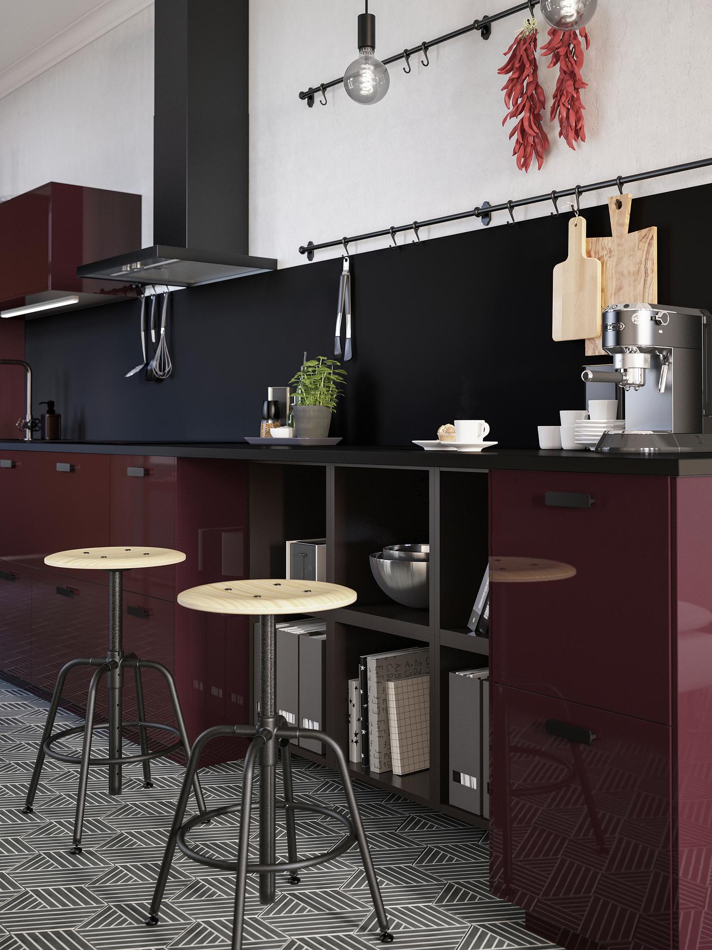 Two stools in pine/black stand by a kitchen countertop in a dark red-brown kitchen. A coffee machine and mugs are on top.