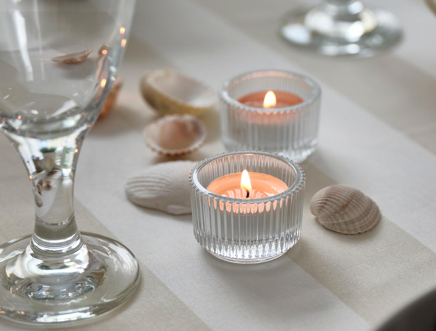 Two tealights in ribbed glass holders with seashells and a wine glass on a softly set table.