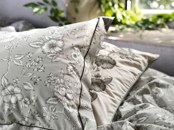 Two versions of IKEA PRAKTBRÄCKA pillowcases in white and grey cotton with vintage flower designs lying on a bed.