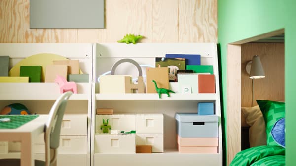 Two white BERGIG book displays with storage, holding a variety of boxes, books and toys, stand against a wall.