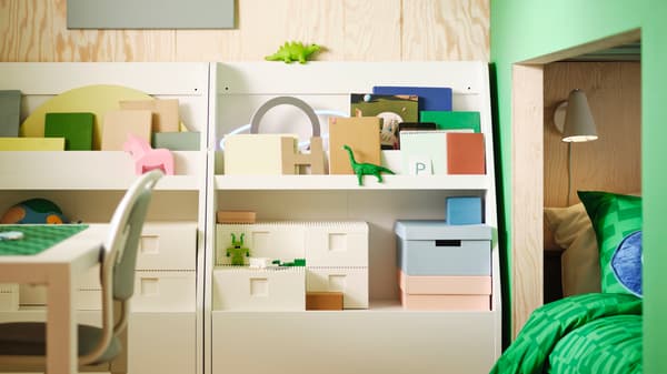 Two white BERGIG book displays with storage, holding a variety of boxes, books and toys, stand against a wall.