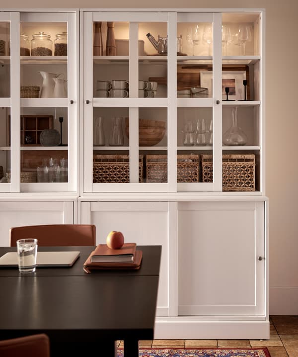 Two white glass-doors cabinets with integrated lighting, storage boxes, decorative items and dinnerware stored inside.