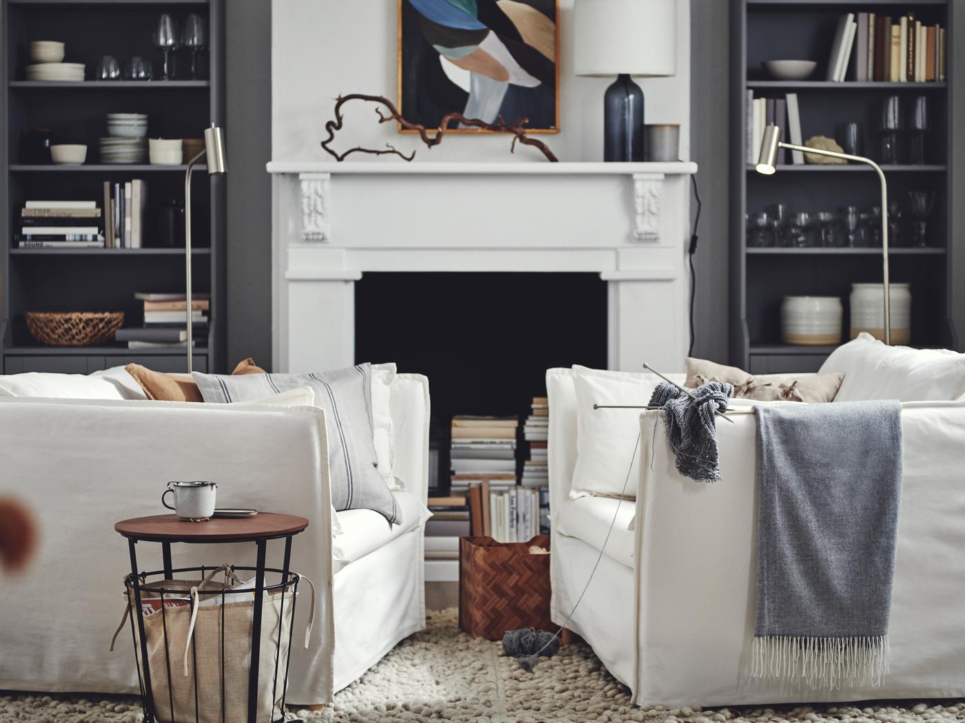 Two white large armchairs facing each other in front of an open fireplace and two floor spotlights, grey bookcases.