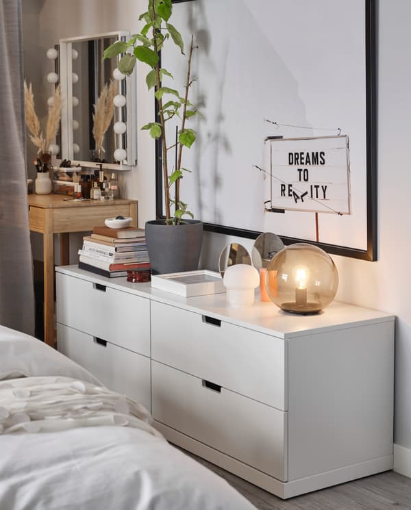 Two white NORDLI chests of drawers stand by a wall. Table lamps, books and decorative items stand on their top surfaces.