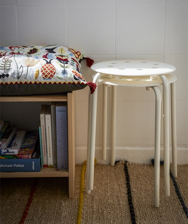 Two white stools stacked next to a bookshelf with a cushion on top.