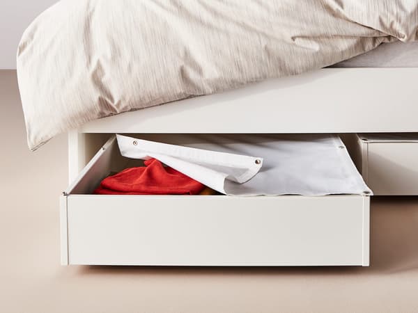 Two white under bed storage boxes, one pulled out with lid partially open, revealing red fabric.