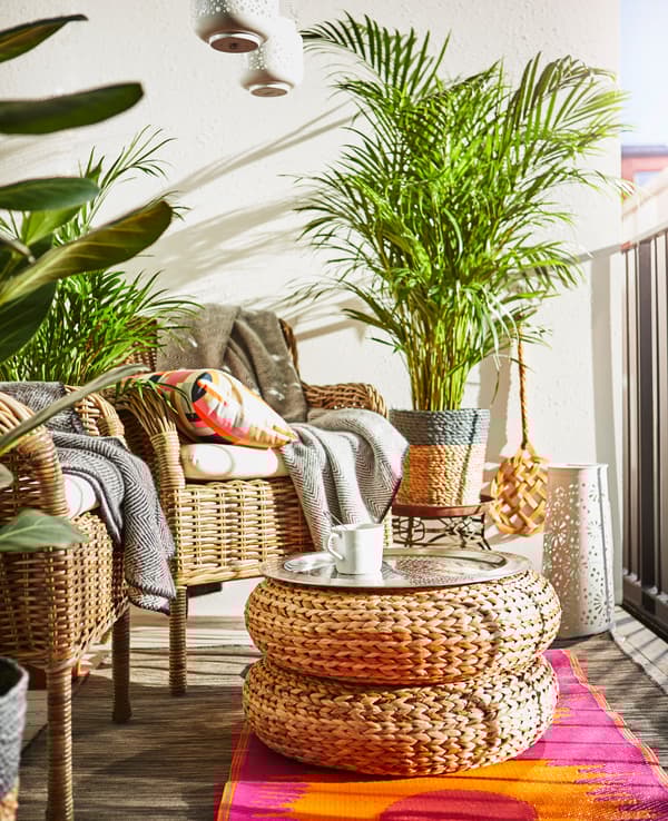 Two wicker armchairs, two stools stacked to make a table, plants and bright rugs sit on a balcony.