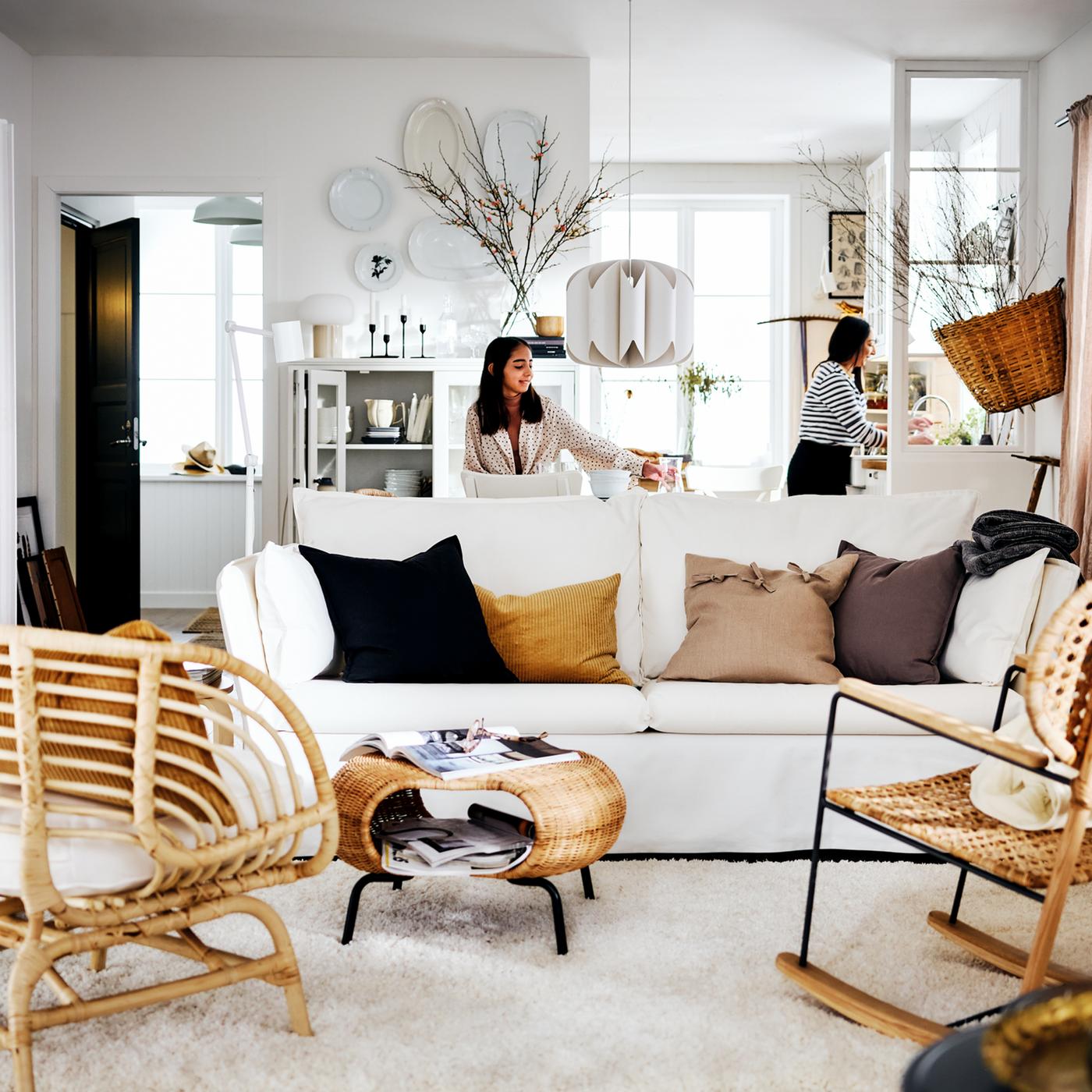 Two women behind a white BACKSÄLEN sofa and a GAMLEHULT ottoman, plus a BUSKBO armchair in rattan on a VOLLERSLEV rug.