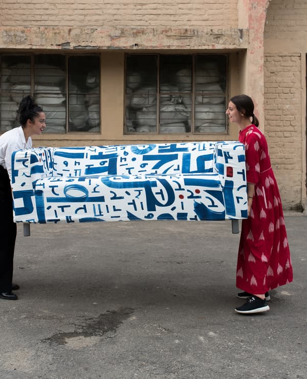Two women holding either end of a sofa with a blue and white textile design on a tarmac surface outside a brick storehouse.