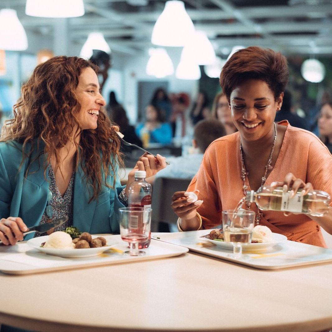 Two women in an IKEA restaurant eating and chatting at a round wooden table