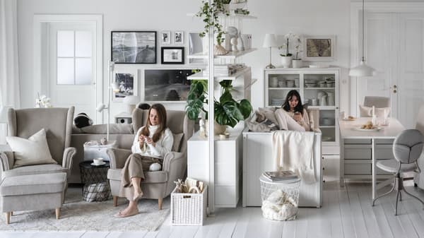 Two women seated, one in a beige STRANDMON armchair, on either side of an ELVARLI storage unit in a large, light living room.