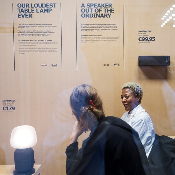Two women sit and talk next to an IKEA SYMFONISK table lamp speaker with graphic posters in the background.