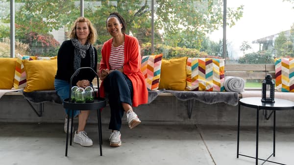 Two women sitting on a bench decorated with colourful pillows and faux furs, smiling into the camera