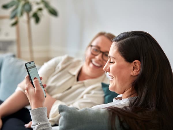 Two women sitting on a sofa smiling, one of them is holding a phone.