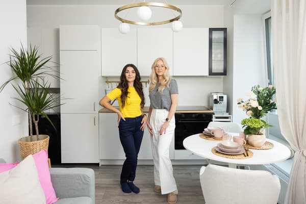 Two women standing in a bright, modern living room with an open-plan kitchen; the space features a round table set with tableware, plants, and contemporary lighting.