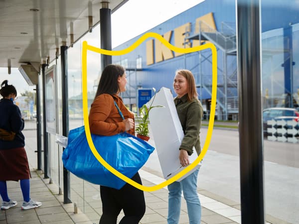 Two women talking outside an IKEA store, one holding a blue FRAKTA bag and the other a white box.