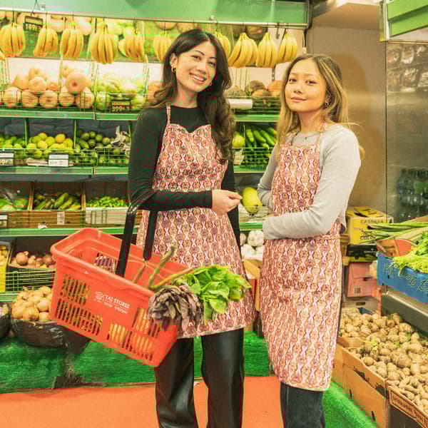 Two women wearing aprons are standing in a shop filled with fruits and vegetables.