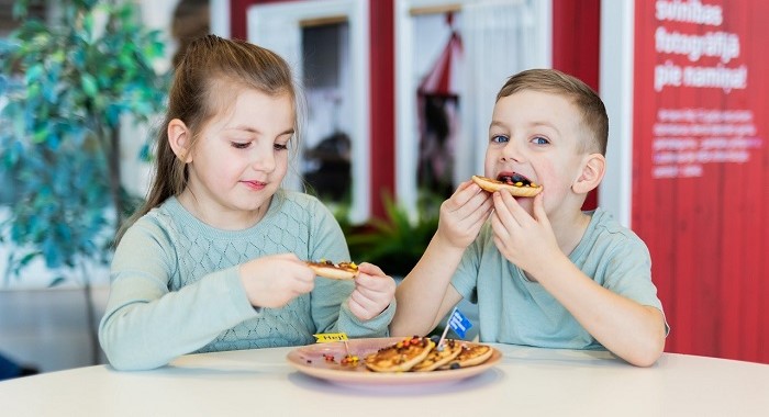 Two young children sitting at a table enjoying small pancakes topped with jam and sprinkles, with a plate full of pancakes between them. The background shows a red wall and a cozy, indoor setting.