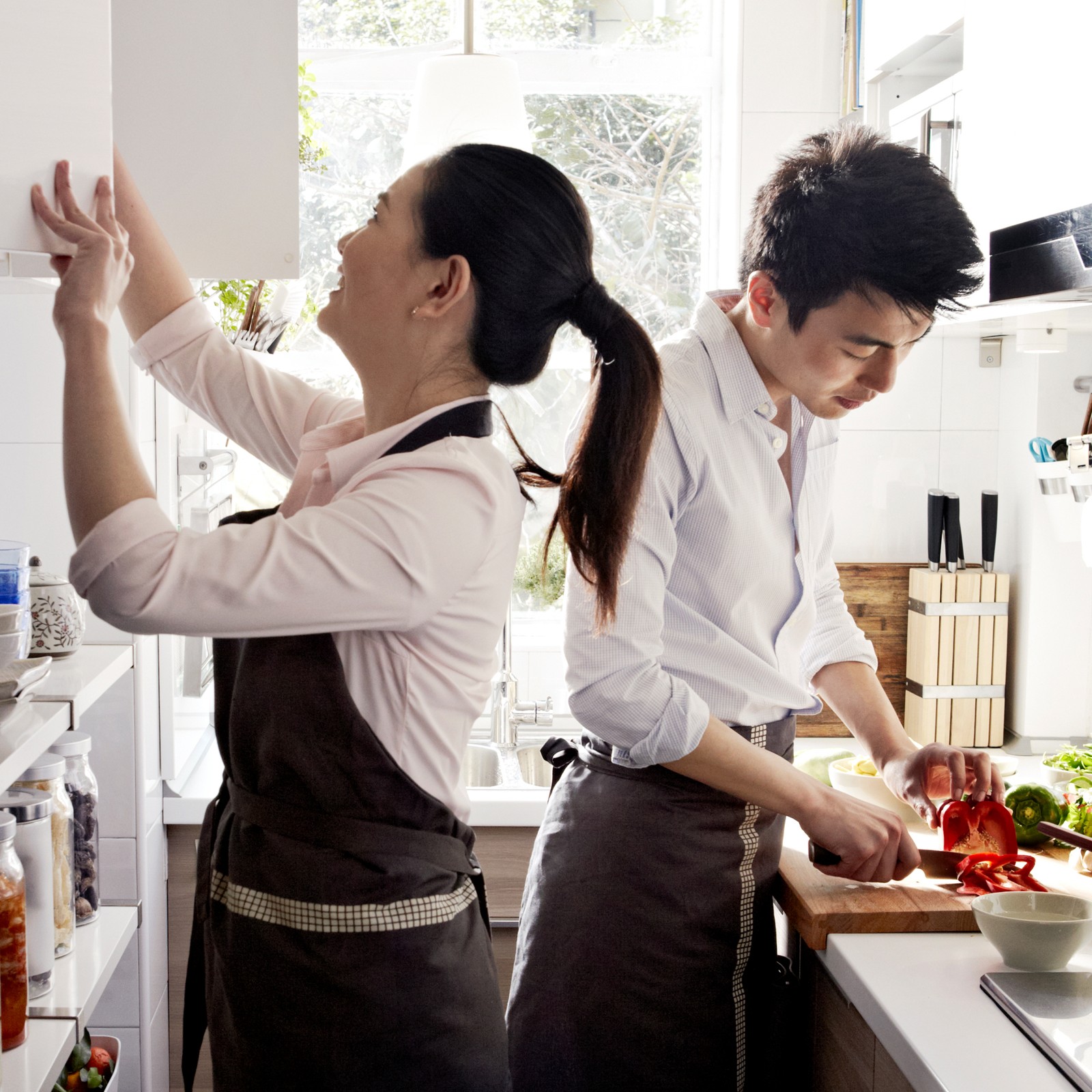 Two young people with aprons in a small space kitchen, one is opening a cabinet and the other is chopping vegetables.