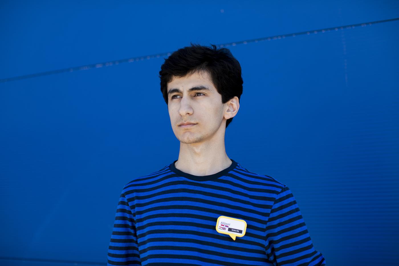 Uchodźca z Tadżykistanu, stoi na tle niebieskiej ściany. Ma na sobie niebieską bluzkę w paski z plakietką z imieniem.  Jest spokojny i poważny, patrzy w dal. 